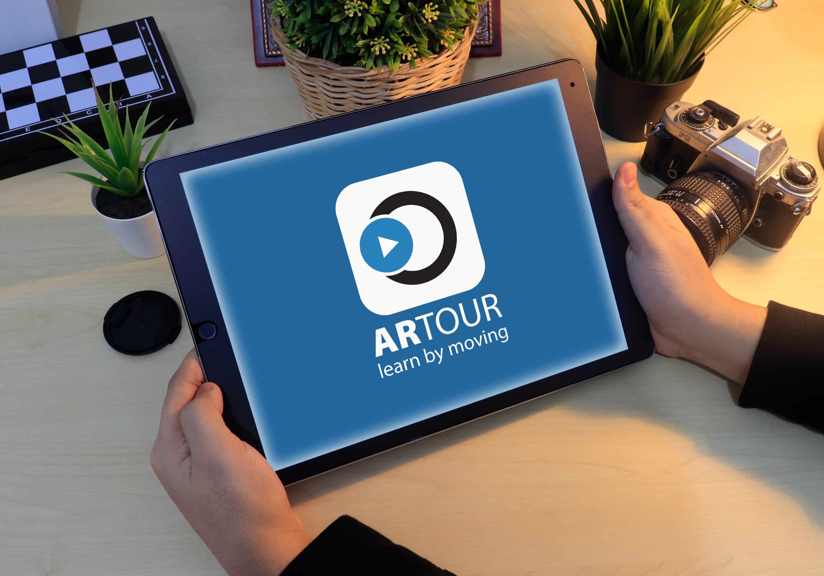 Roke drzijo tablico, ki prikazuje logotip aplikacije ARTour learn by moving na leseni mizi ob fotoaparatu.