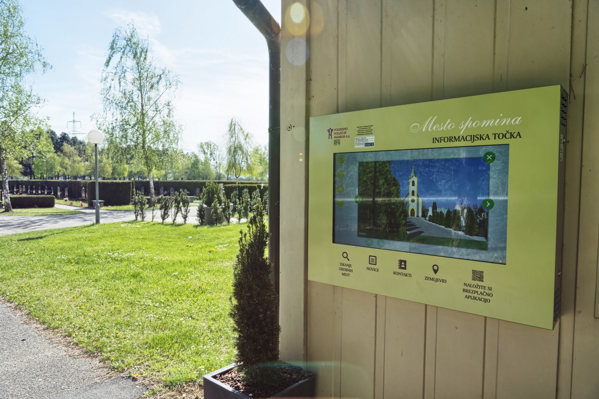 Interaktivna digitalna informacijska točka Mesto spomina na leseni steni ob urejenem parku.