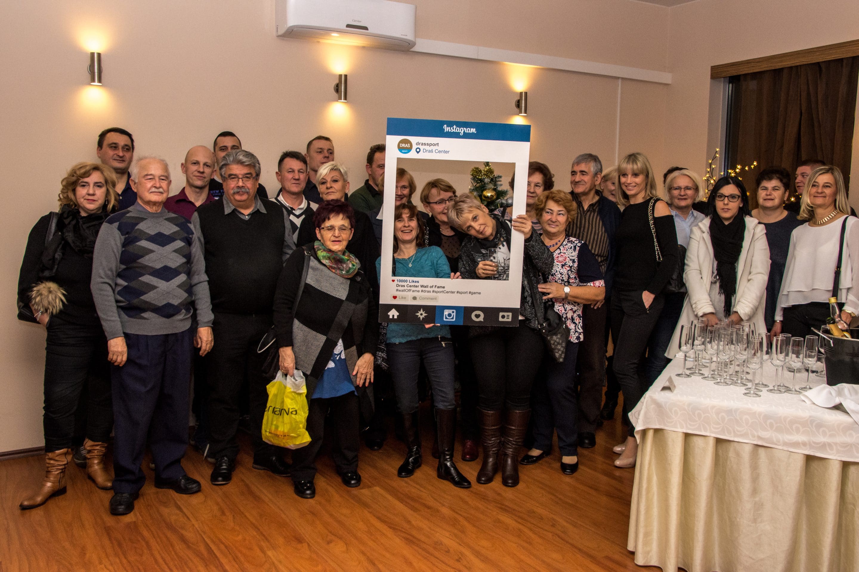 Skupina ljudi na dogodku pozira z velikim kartonastim okvirjem za Instagram ob obloženi mizi.