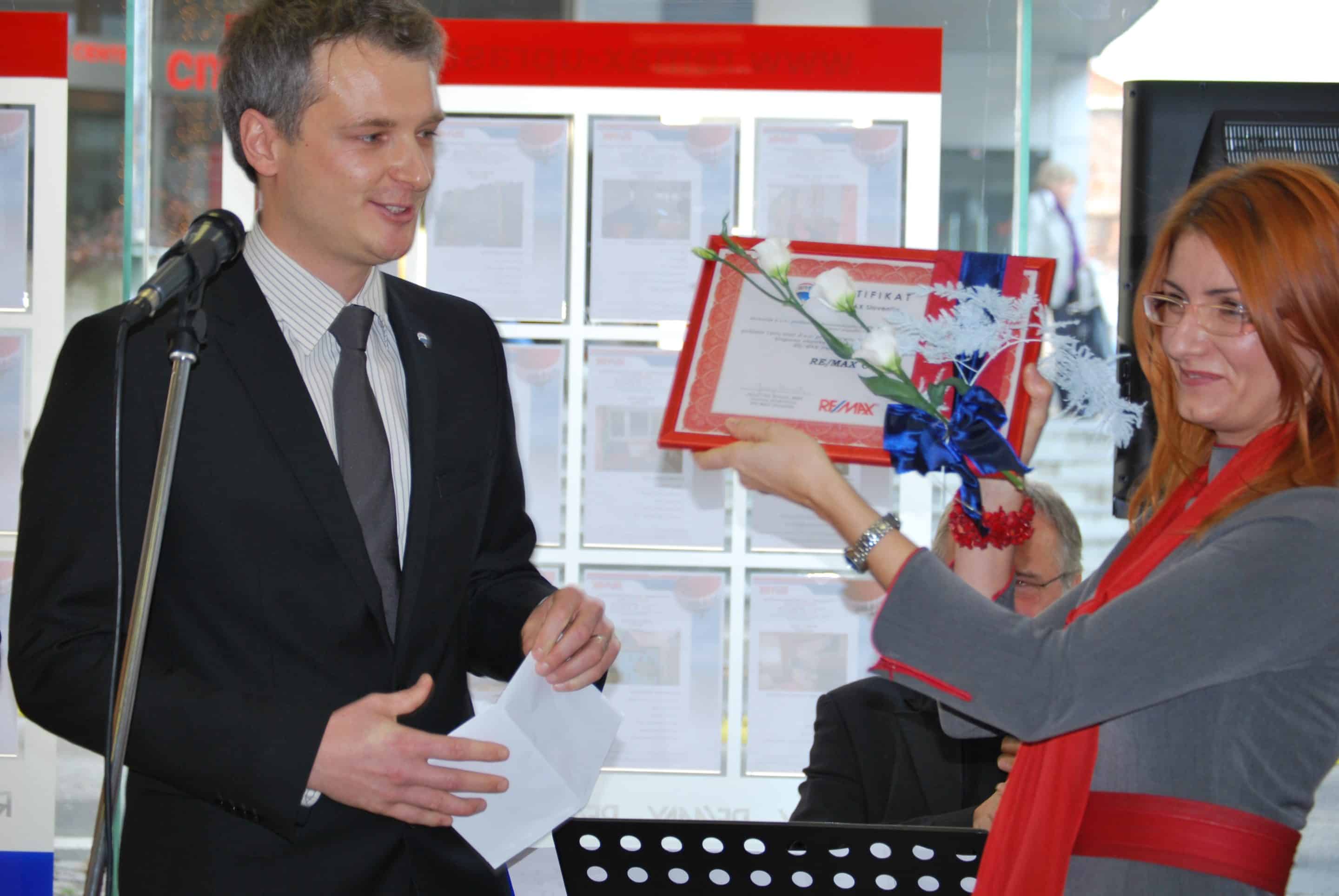 Ženska v sivi obleki podeljuje uokvirjeno priznanje in cvet moškemu v obleki pred mikrofonom.