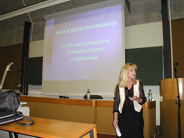 Svetlolasa predavateljica pred projekcijskim platnom z naslovom Vedenje kupcev in porabnikov v predavalnici.