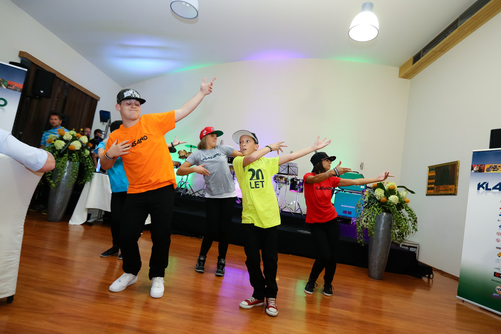 Mladi hip-hop plesalci v pisanih majicah izvajajo koreografijo na prireditvi v dvorani.