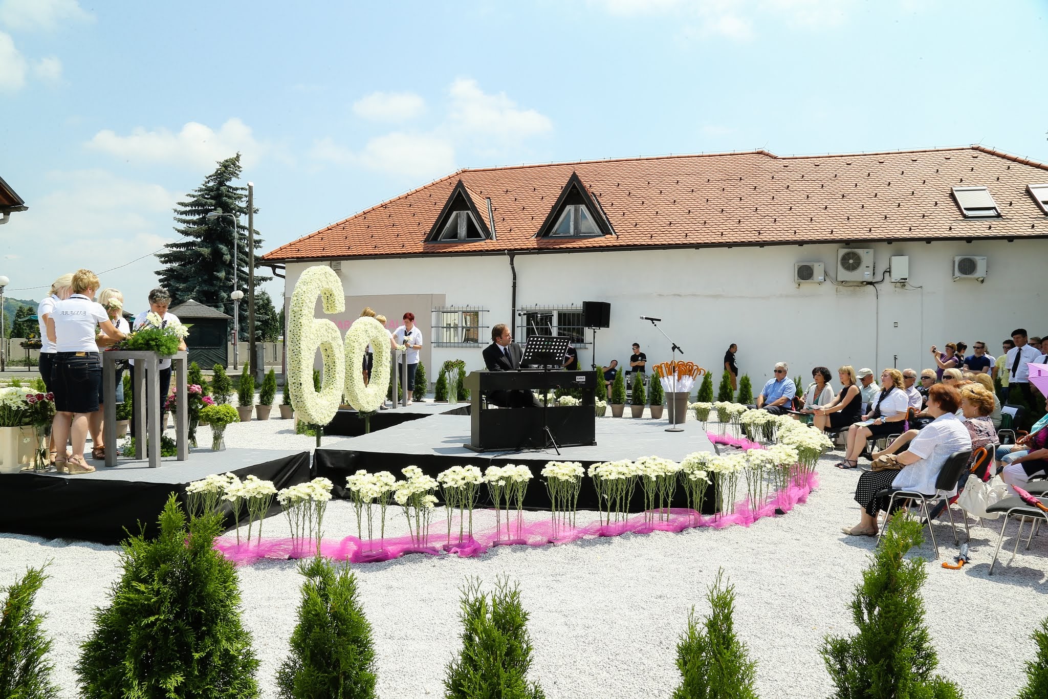 Praznovanje 60. obletnice na prostem s pianistom na odru, cvetlično dekoracijo in občinstvom pred stavbo.