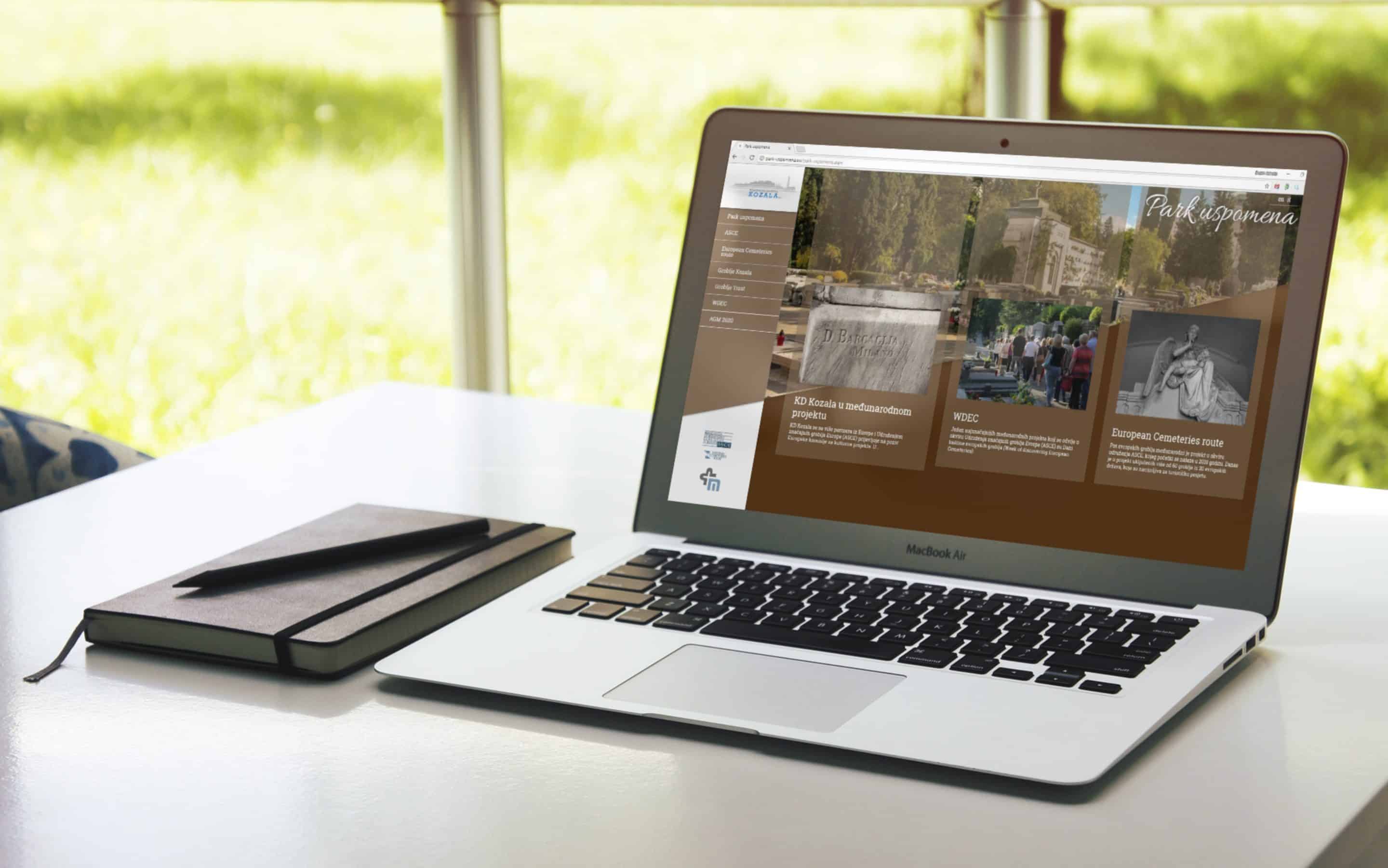 Prenosni računalnik s spletno stranjo Park uspomena in beležnica na beli mizi z zelenim ozadjem.