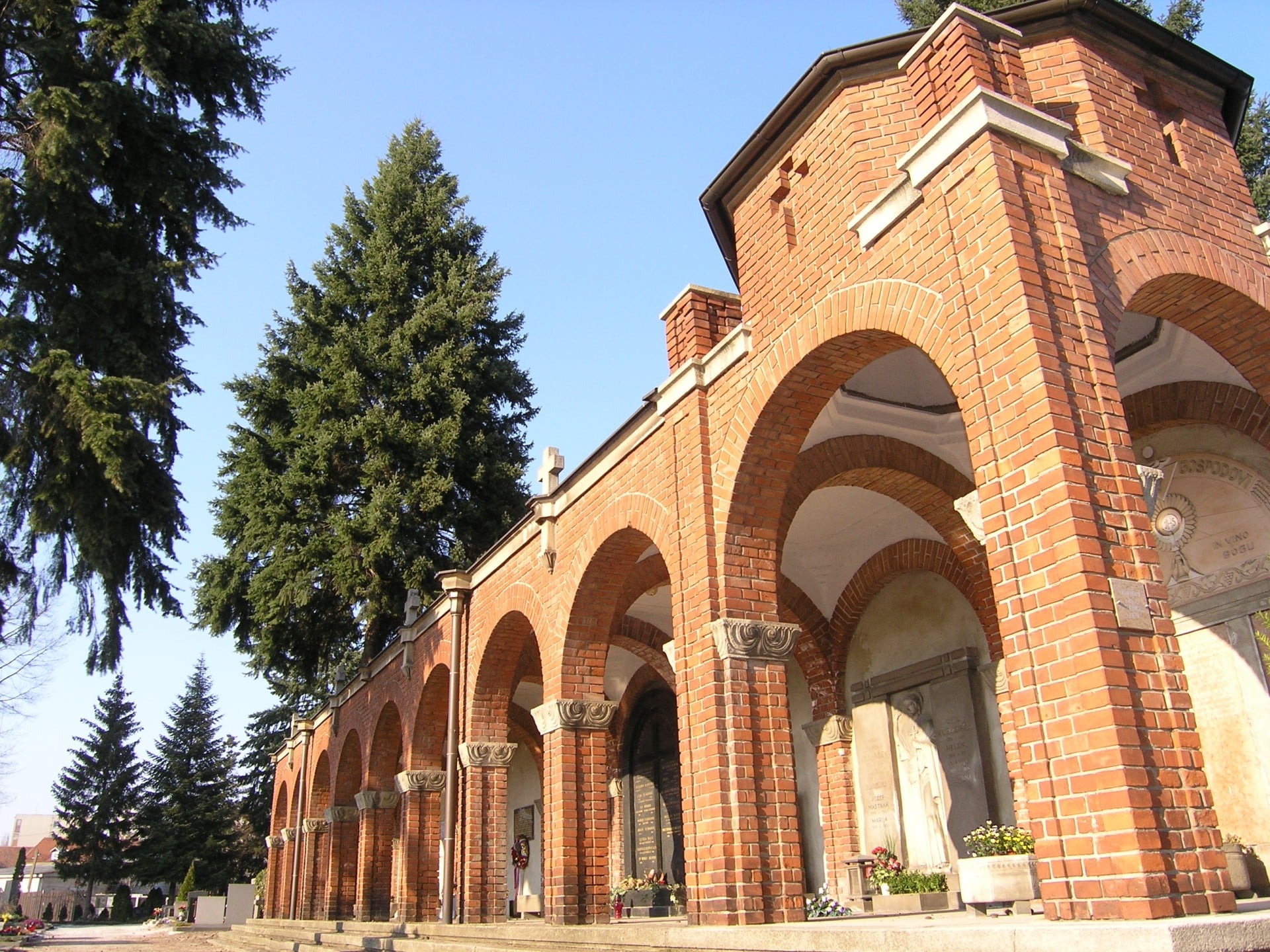 Rdeče opečne arkade na pokopališču Pobrežje v Mariboru pod jasnim nebom z visokimi smrekami.