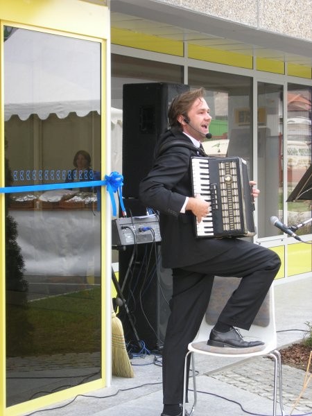 Moški v črni obleki igra na harmoniko in poje na otvoritvenem dogodku pred vhodom z modrim trakom.