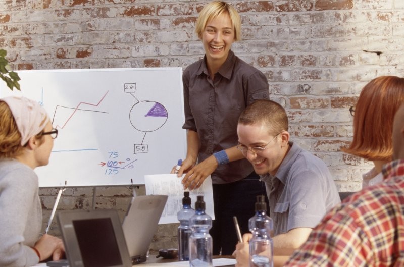 Nasmejana predavateljica ob tabli z grafi vodi sestanek za strategijo marketinga.