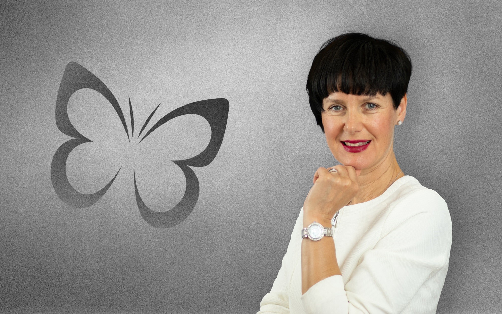marketing-agencija-kainoto-natalija-popovic-1 Nasmejana Natalija Popovič v beli bluzi ob stiliziranem logotipu metulja na sivi podlagi.