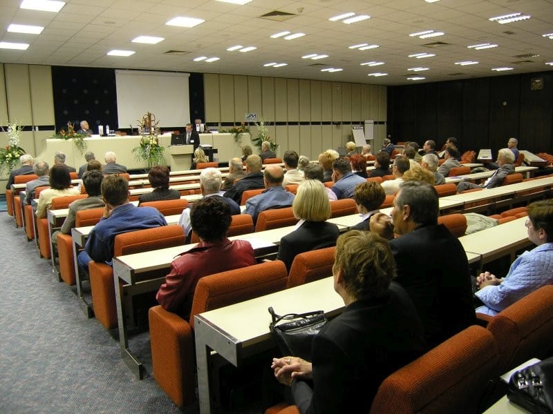 Udeleženci simpozija sedijo v vrstah v veliki predavalnici in poslušajo predavatelje na odru.