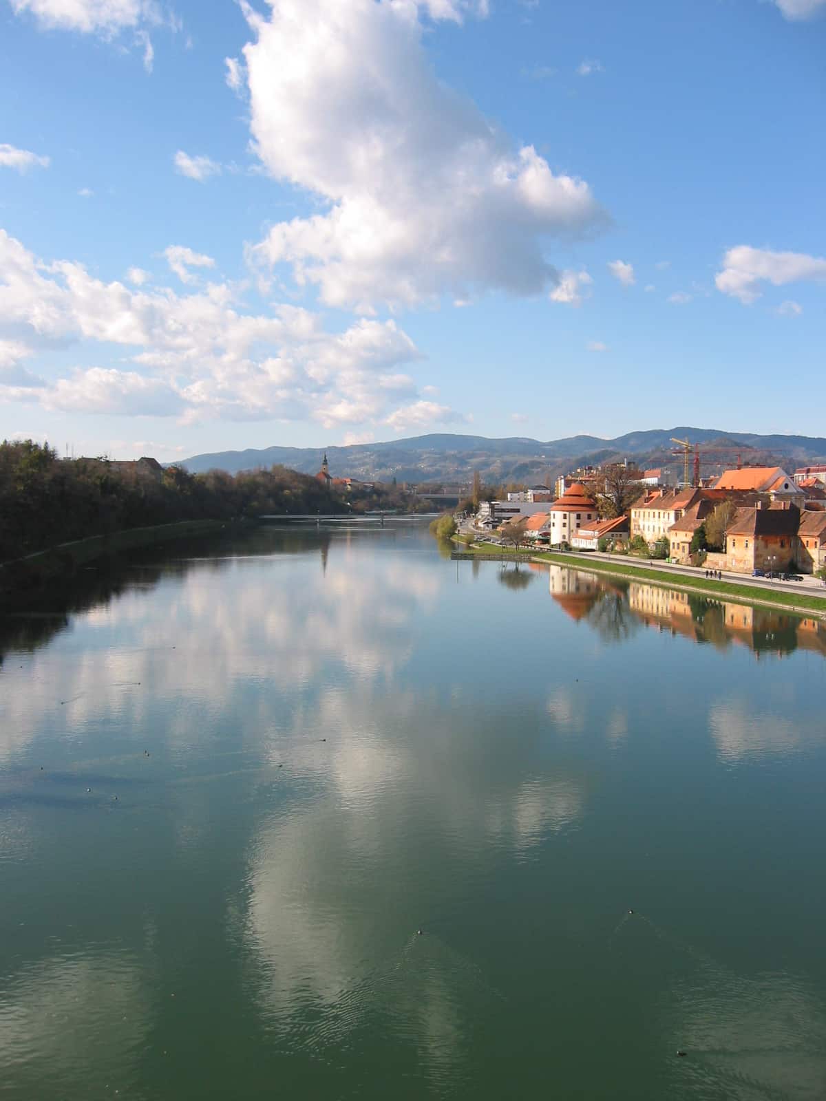 Reka Drava v Mariboru z odsevom oblakov na gladini in zgodovinskim Lentom na desnem bregu.