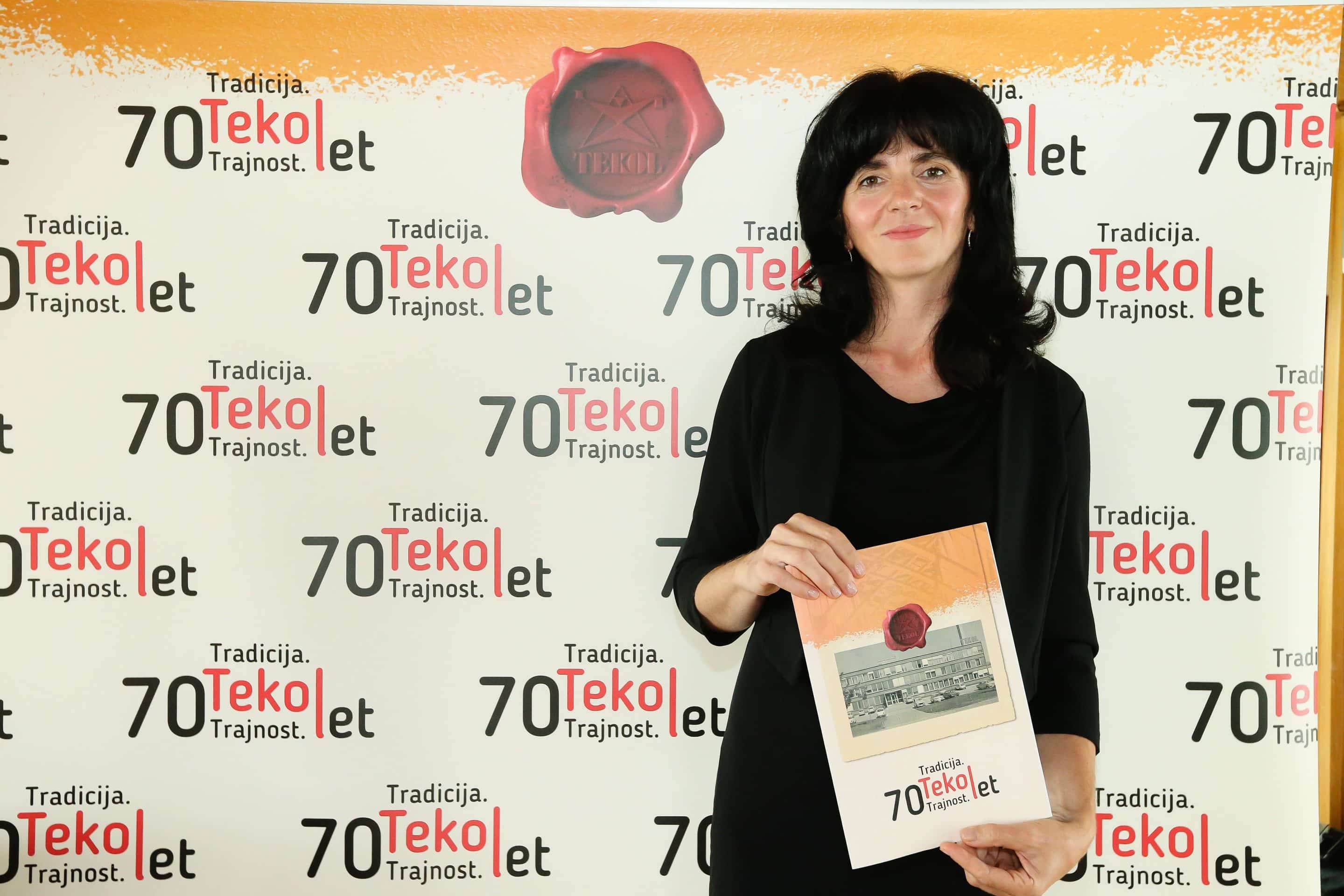 Ženska s predstavitveno brošuro pred steno z logotipi za 70-letnico podjetja Tekol.