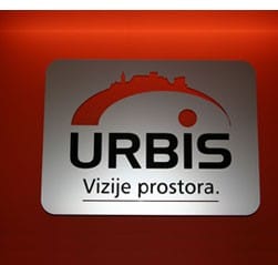 Logotip podjetja Urbis na kovinski plošči z rdečo silhueto mesta in sloganom Vizije prostora na rdeči steni.