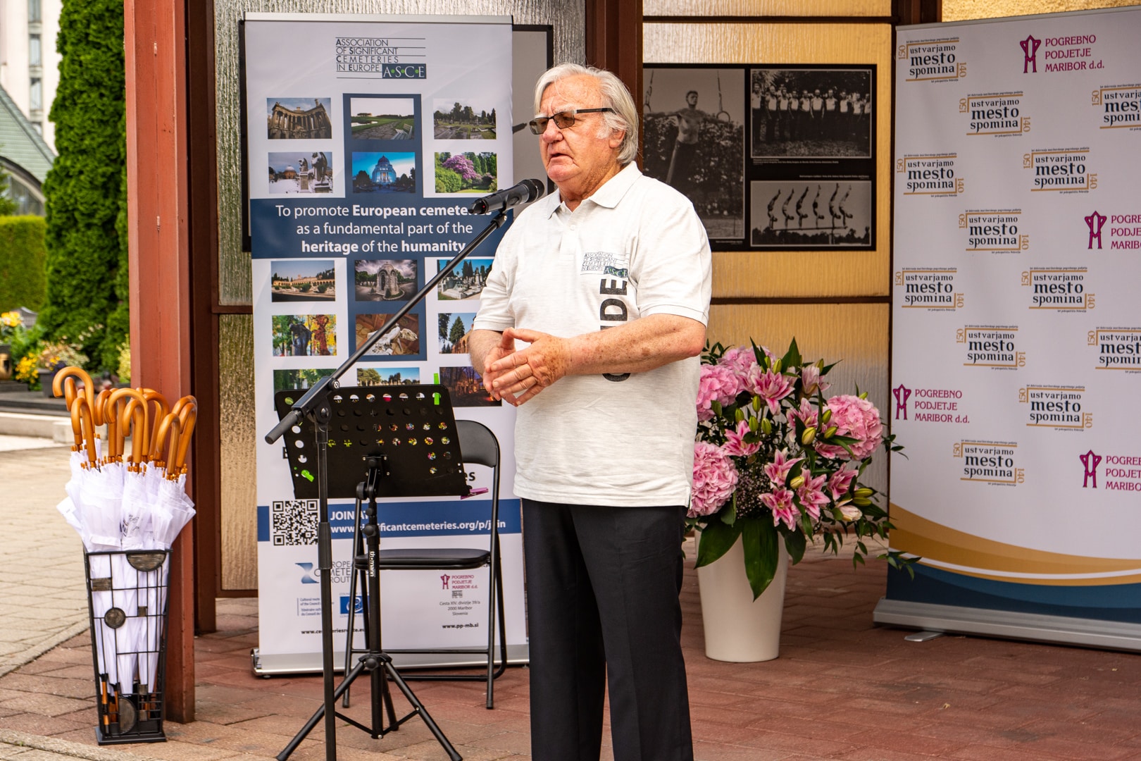 Starejši moški z očali govori v mikrofon na dogodku pred promocijskimi panoji ASCE in Pogrebnega podjetja Maribor.