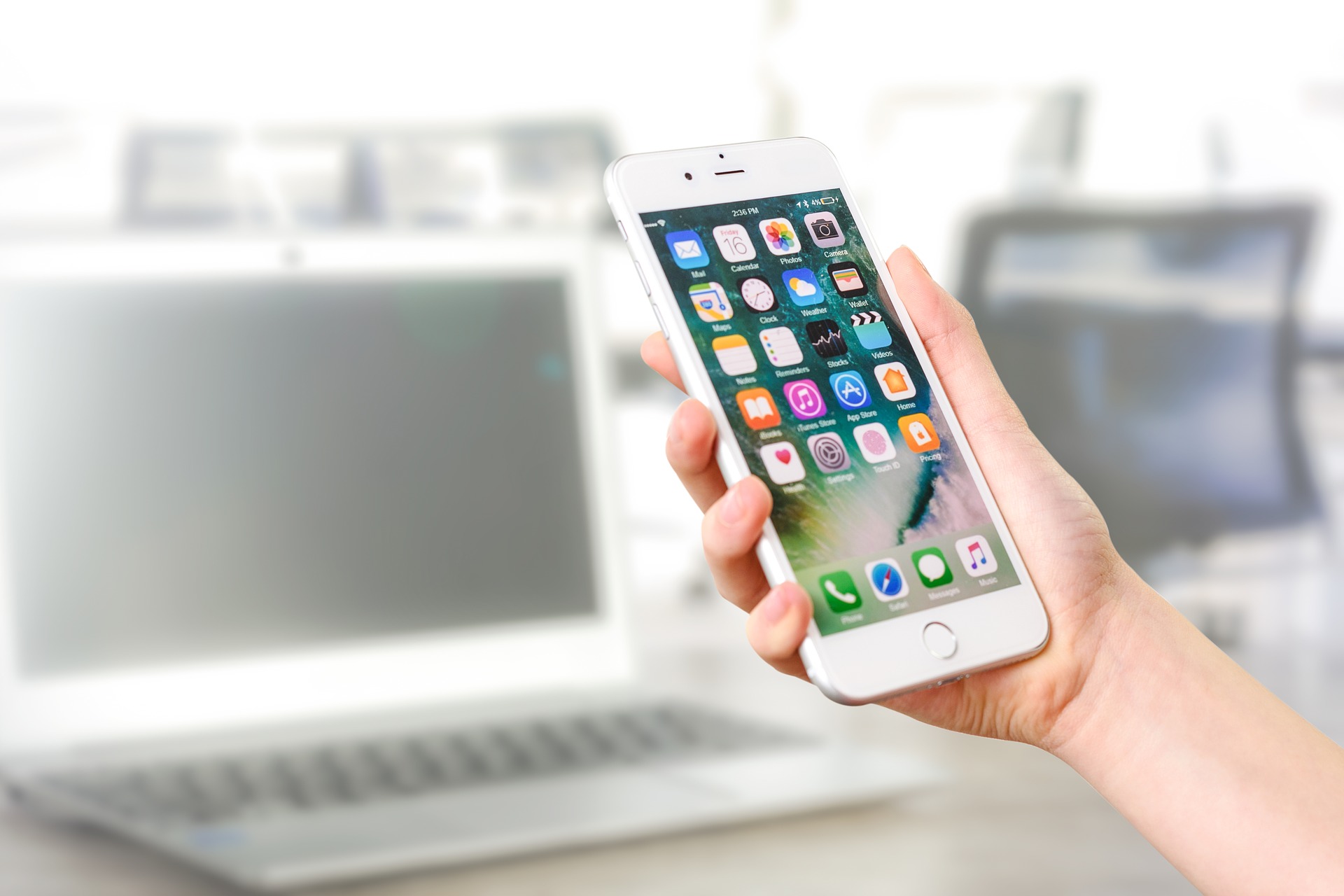 Roka, ki drži bel pametni telefon s prikazanimi ikonami mobilnih aplikacij, v ozadju pa je zamegljen prenosnik.
